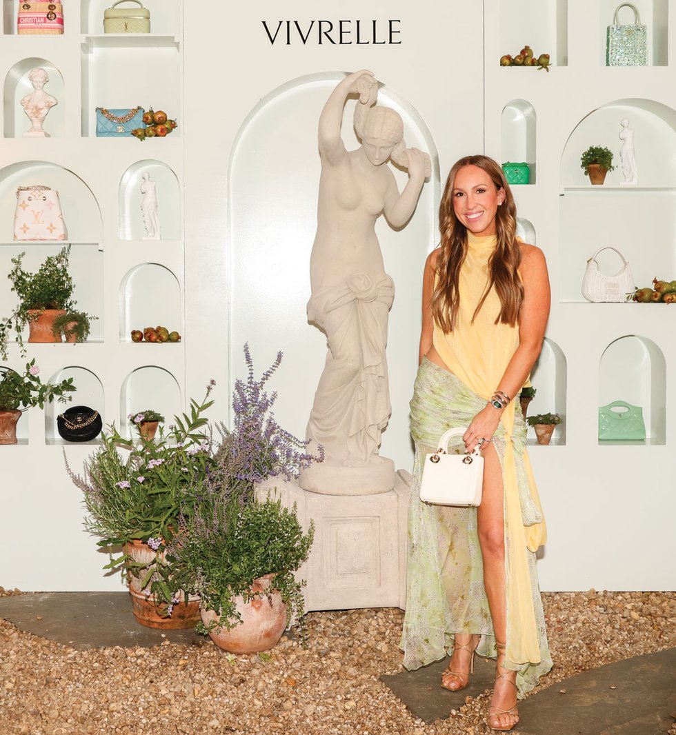 Blake Geffen in a yellow top and patterned skirt beside a statue and a Vivrelle accessories display