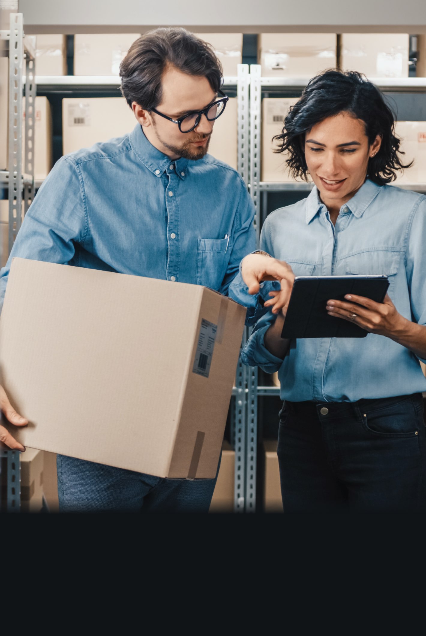Two people consult a tablet among warehouse shelves