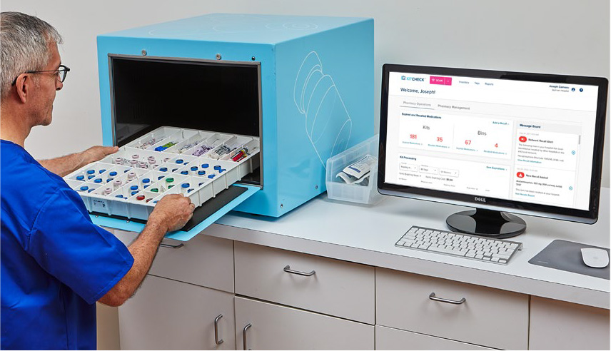 A pharmacy technician loads a medication tray into a blue KitCheck scanning station beside its software dashboard