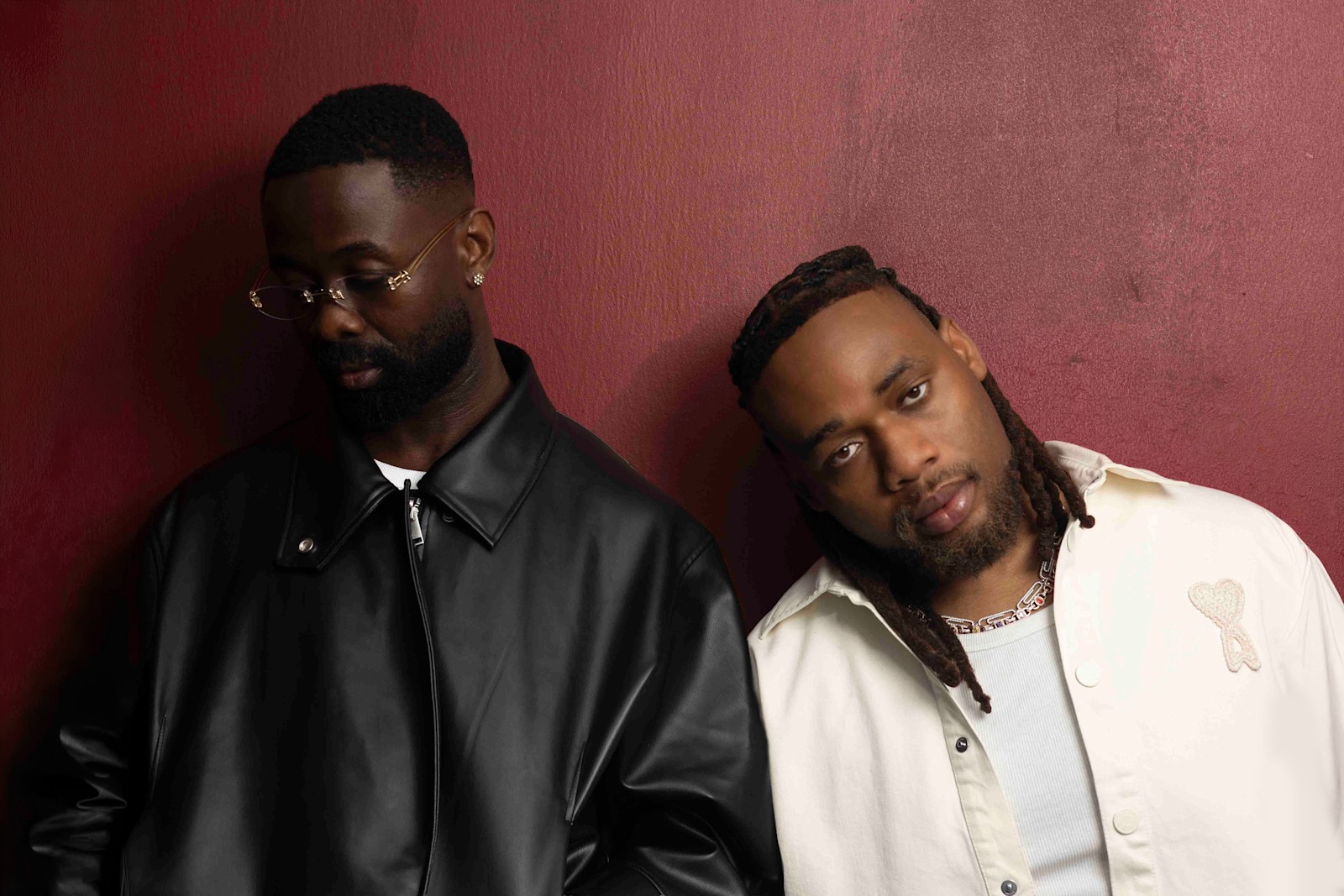 Producer Sarz in a black leather jacket beside BNXN in a cream jacket against a red wall