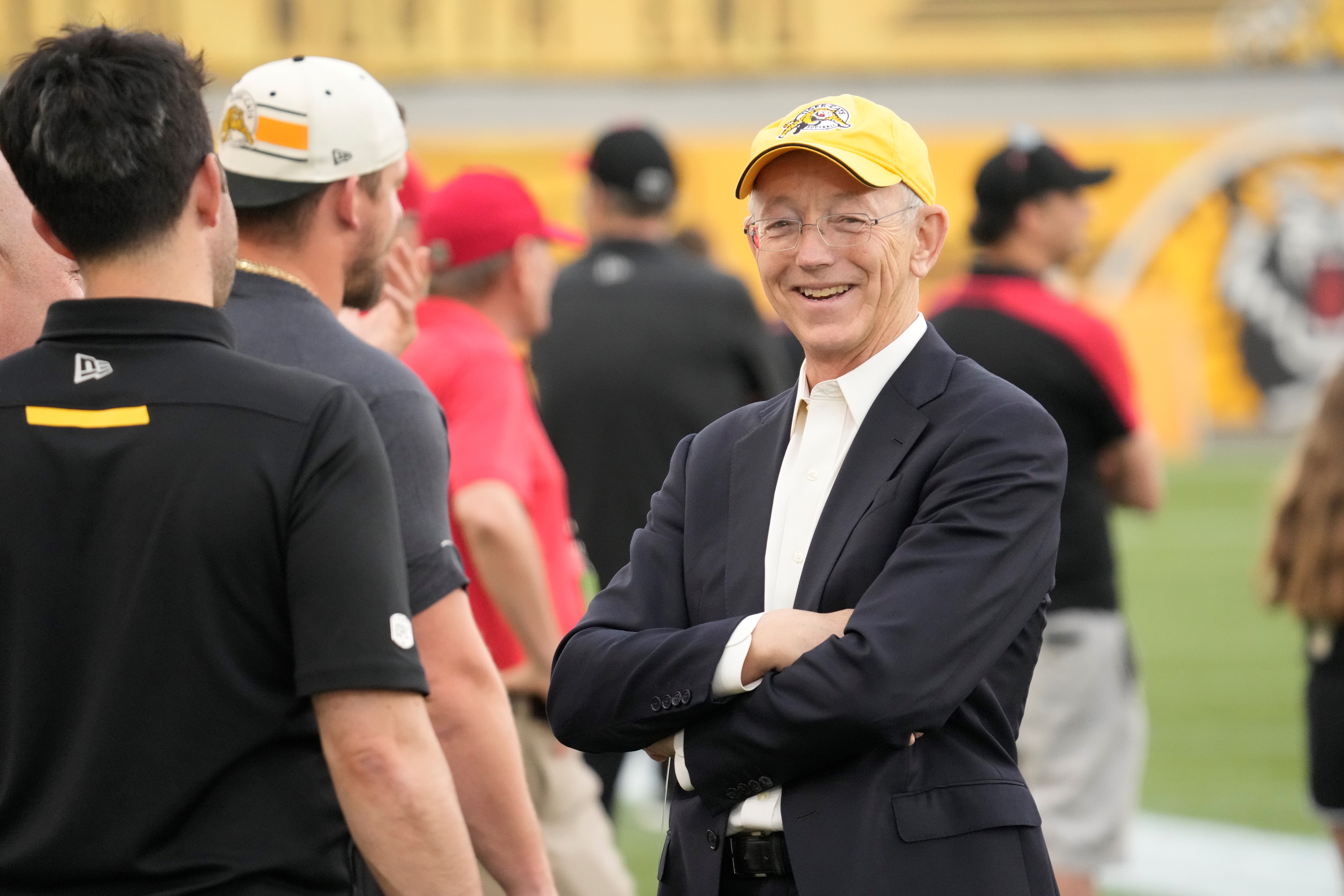 Bob Young in a yellow Tiger-Cats cap and dark jacket, smiling among people on a football field