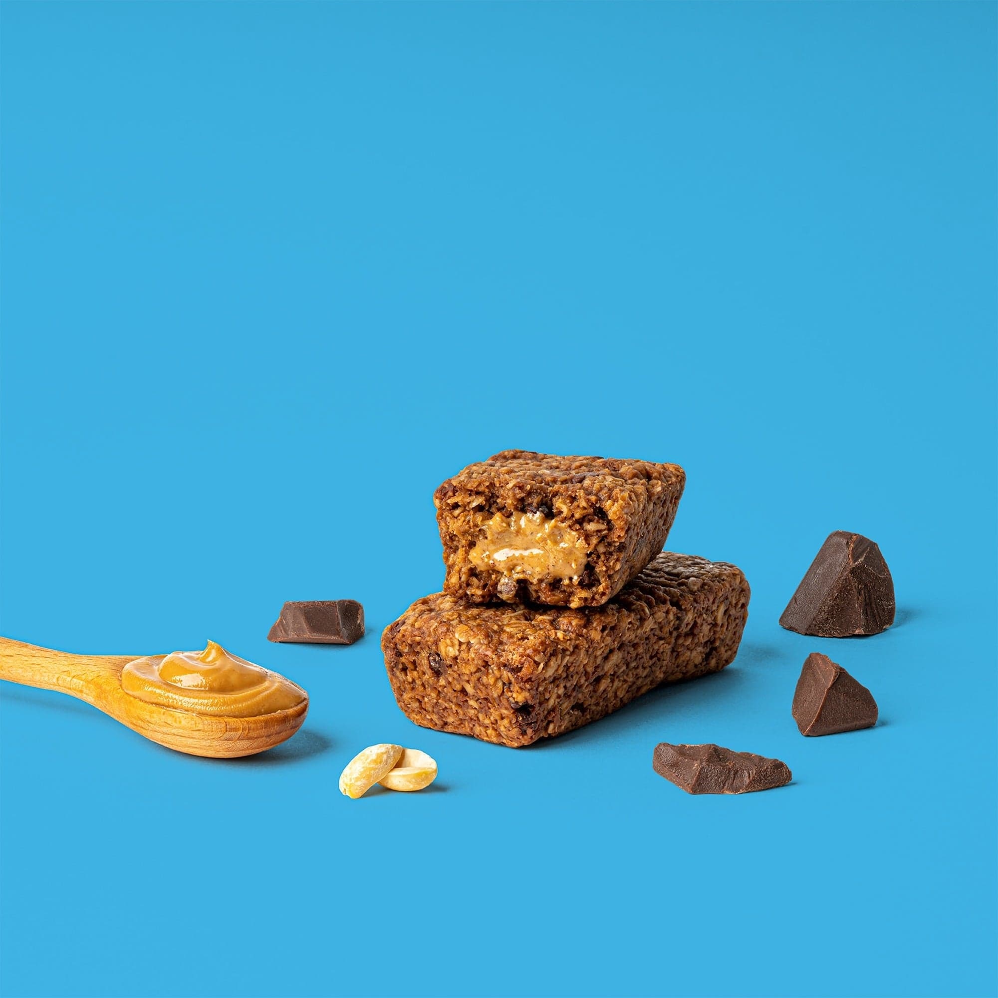A peanut-butter-filled chocolate oat bar cut open on a blue background, with peanuts and chocolate nearby
