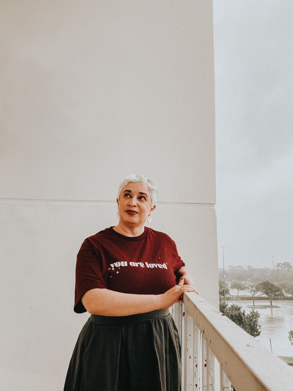 A person wears a burgundy You Are Loved shirt beside a balcony railing.