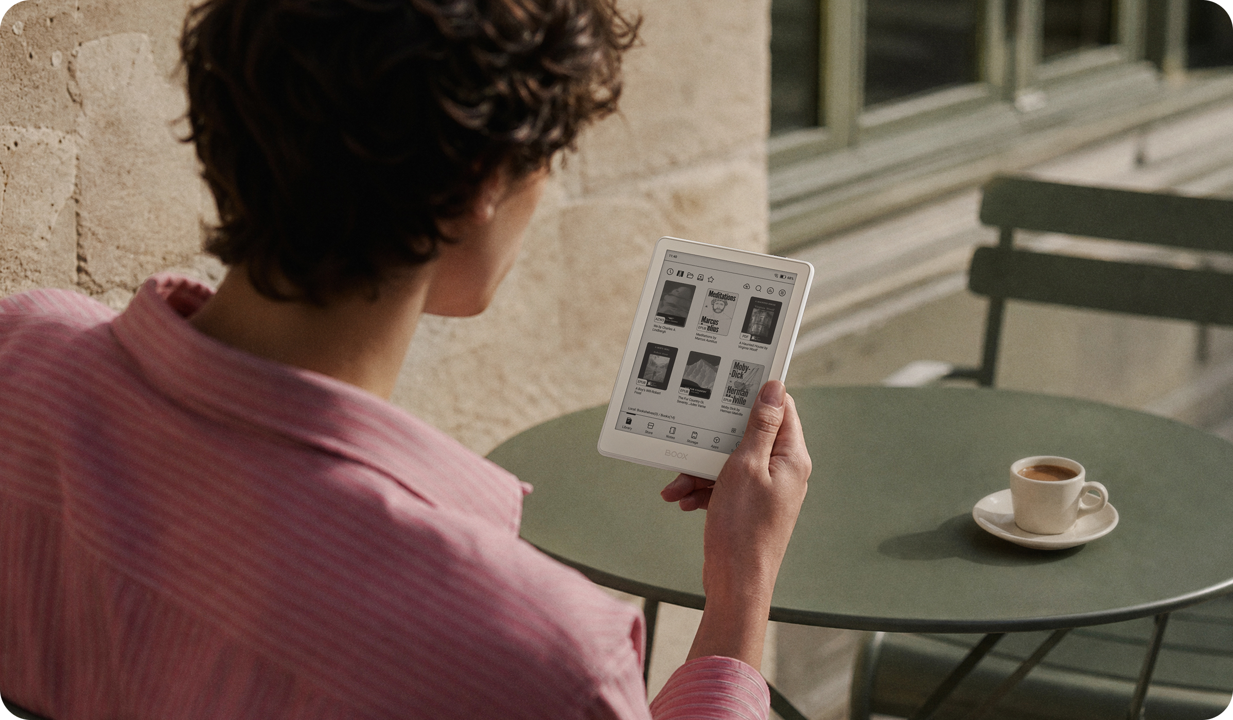 Over-the-shoulder view of the BOOX Go 6 Gen II showing its library grid of ebooks, held at a cafe table with an espresso