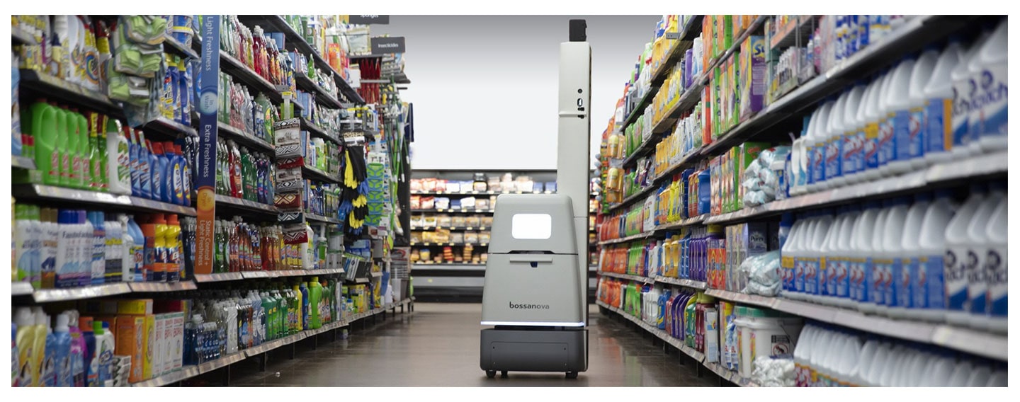 A tall white Bossa Nova robot beside supermarket shelves of household products