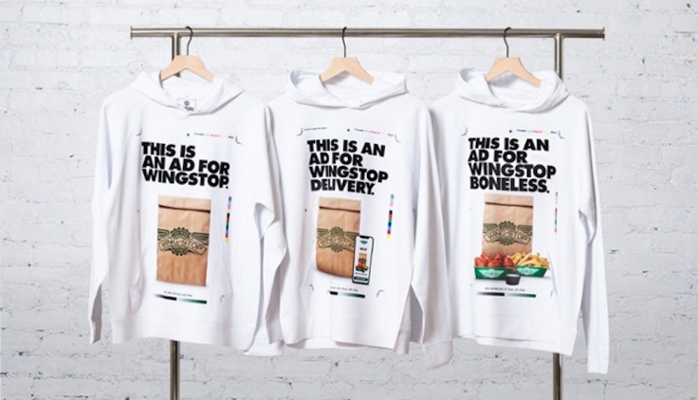 Three white Wingstop advertising hoodies hanging against a brick wall