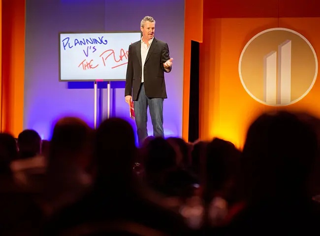 Brad Sugars speaking to an audience beside a handwritten planning board