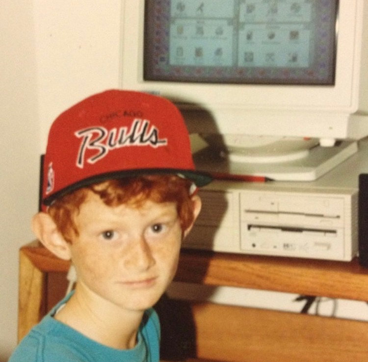 A young Bradley DeLoatche in a Chicago Bulls cap sitting beside an early home computer