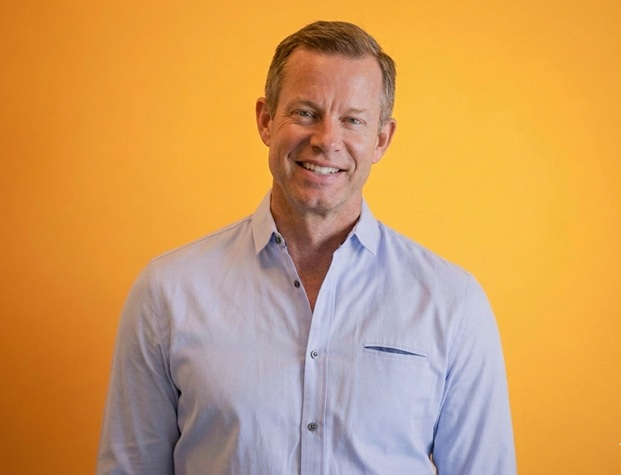 Brett Carlson wearing a pale blue shirt against a yellow background
