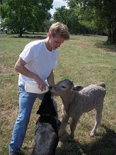 A younger Brett Goodson bottle-feeding a calf outdoors, with a black dog beside him