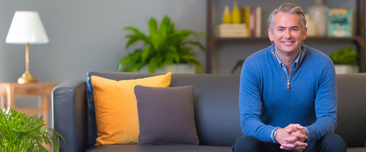 Brian Doubles seated on a sofa, wearing a blue sweater