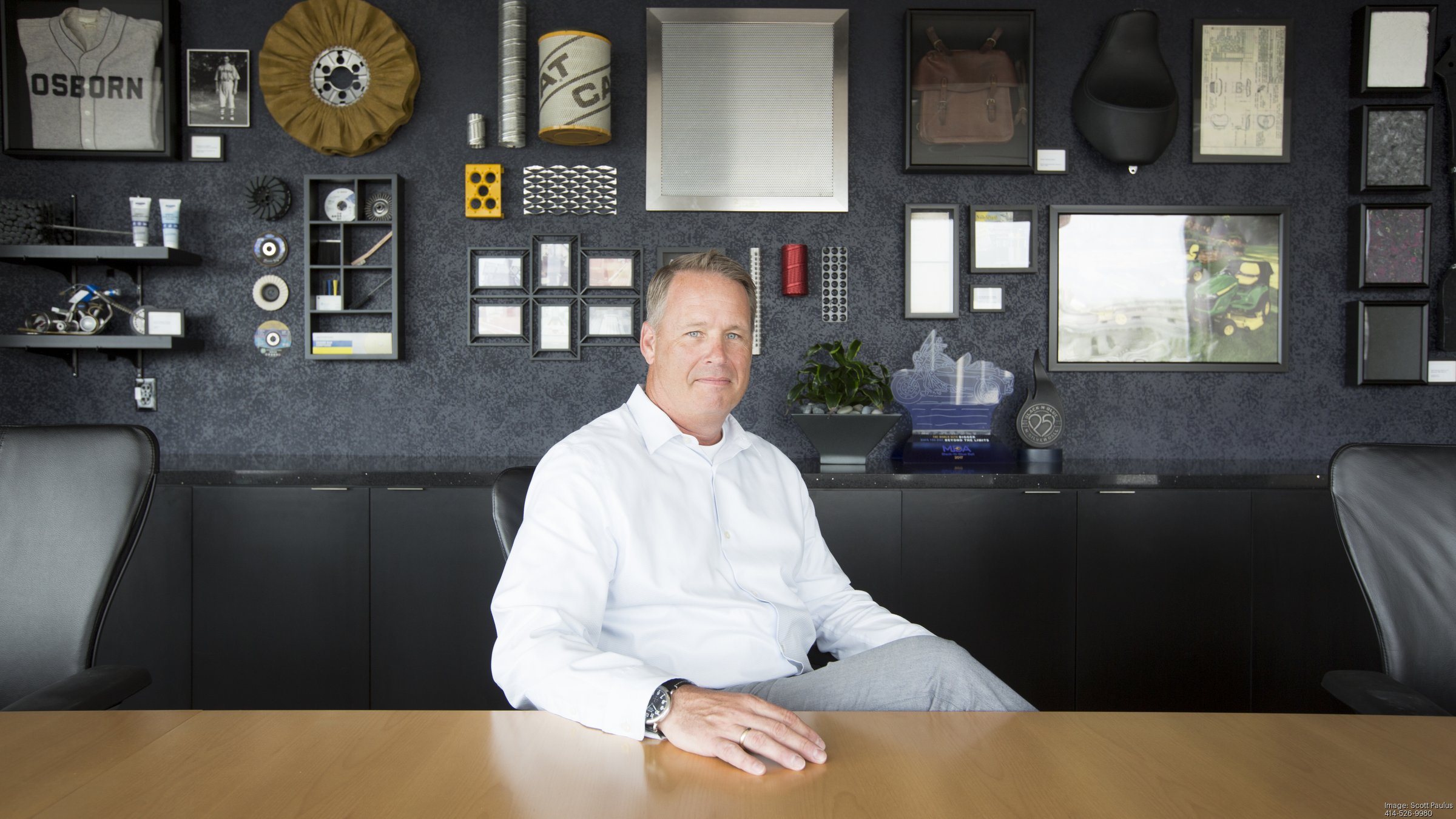Brian Kobylinski seated at a conference table, with industrial products and company memorabilia displayed behind him.
