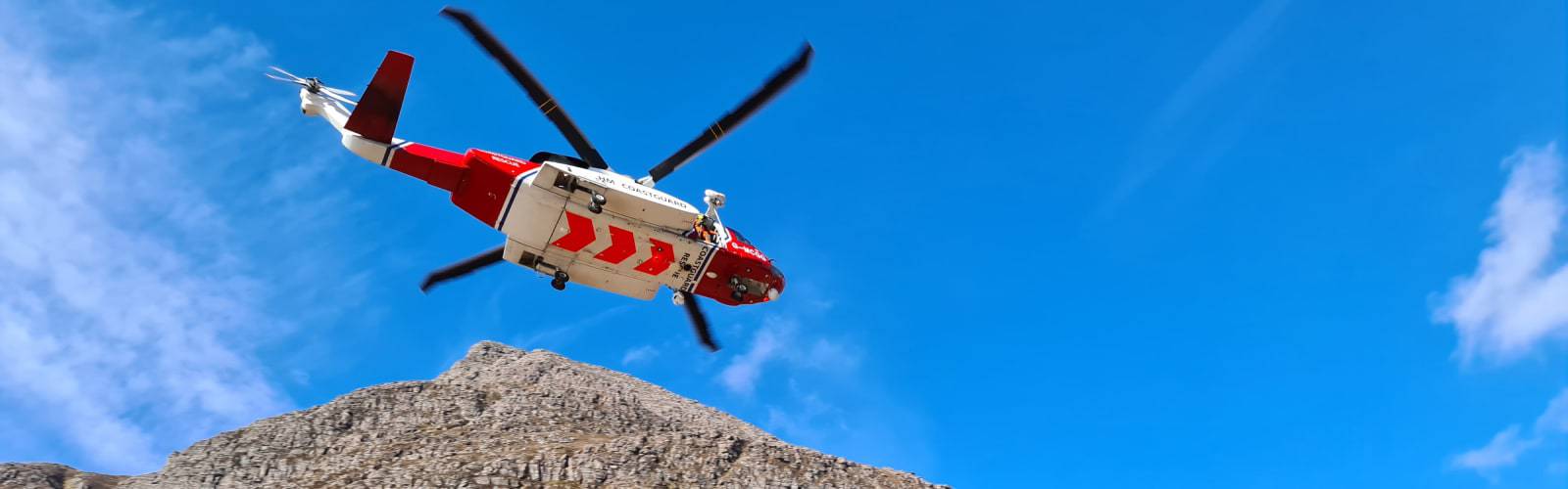 Red and white HM Coastguard helicopter flying above rocky terrain beneath a blue sky