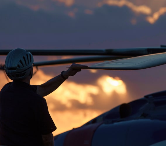 A helmeted crew member beside a helicopter rotor blade at sunset
