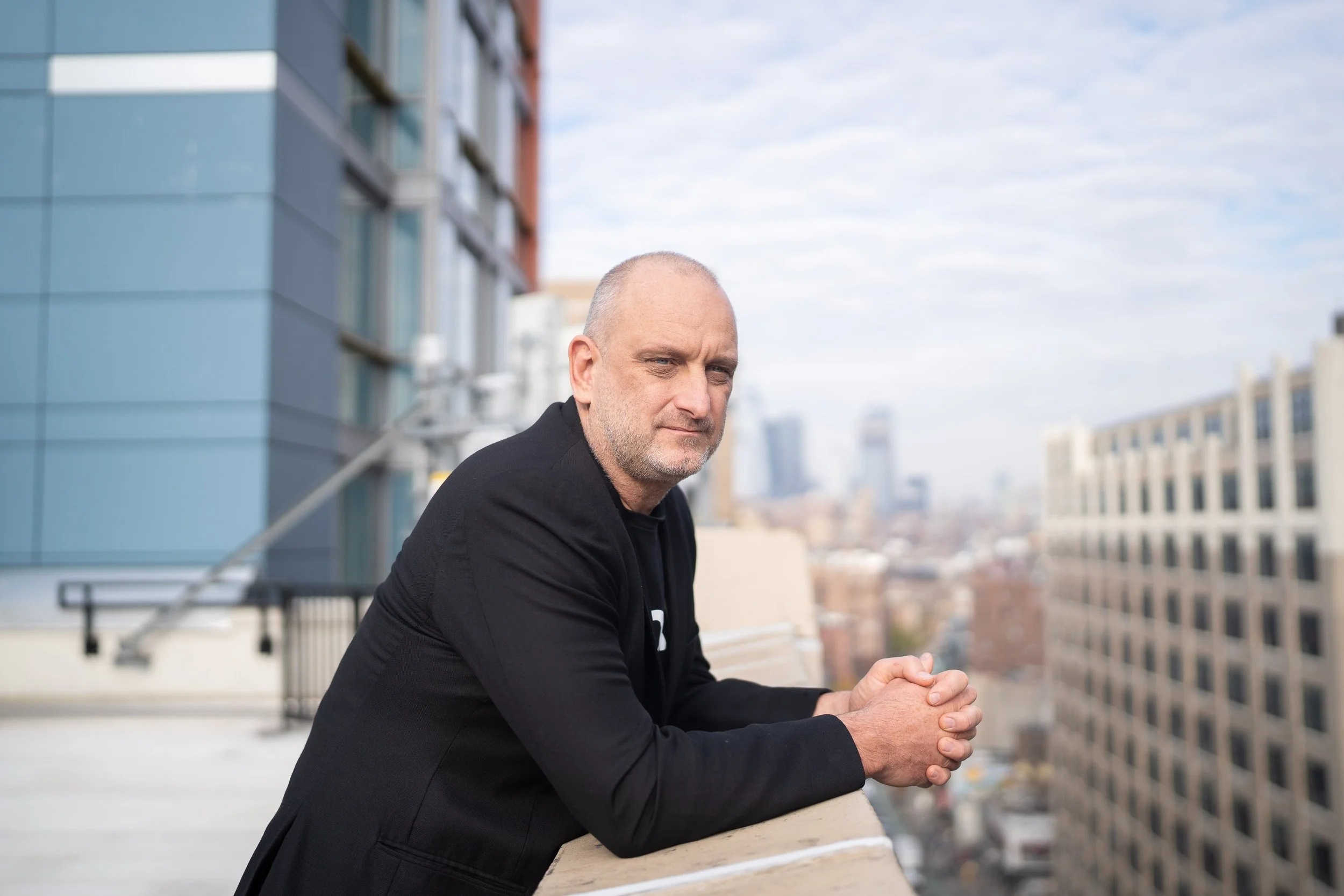 Bruce Buchanan leaning on an outdoor balcony railing, with city buildings behind him