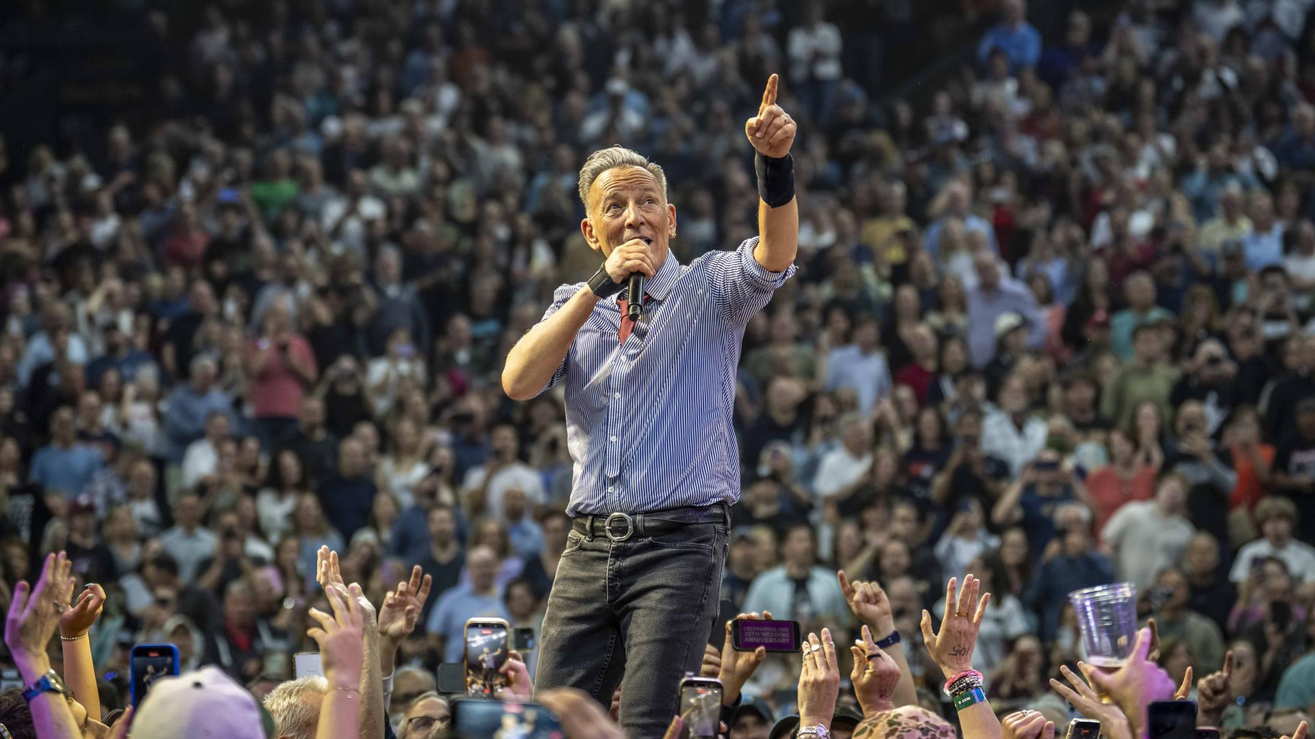 Bruce Springsteen standing among a concert crowd with a microphone, pointing upward