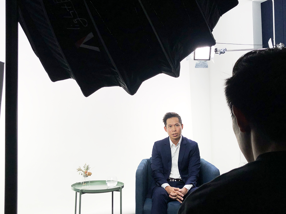 Bryan Koh seated during a filmed interview, with the interviewer and studio lighting visible