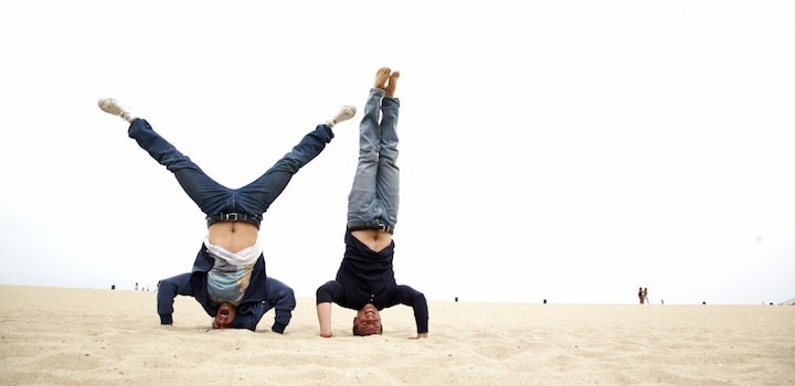 Two people in suits doing headstands side by side on a beach, in a photograph accompanying Bryce Maddock’s 2015 interview