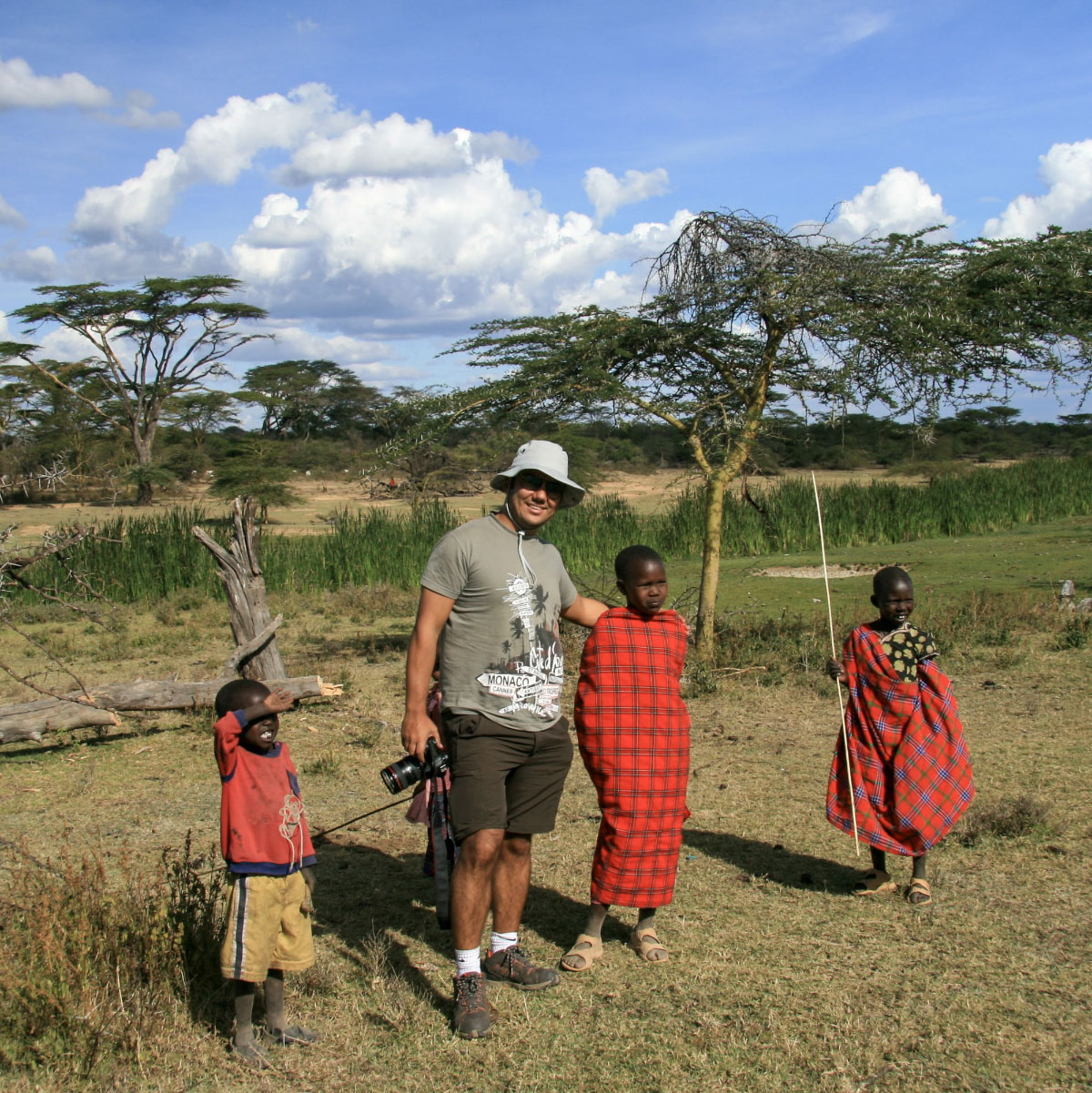 Burak Arik with a camera beside three children in a grassy landscape in Kenya
