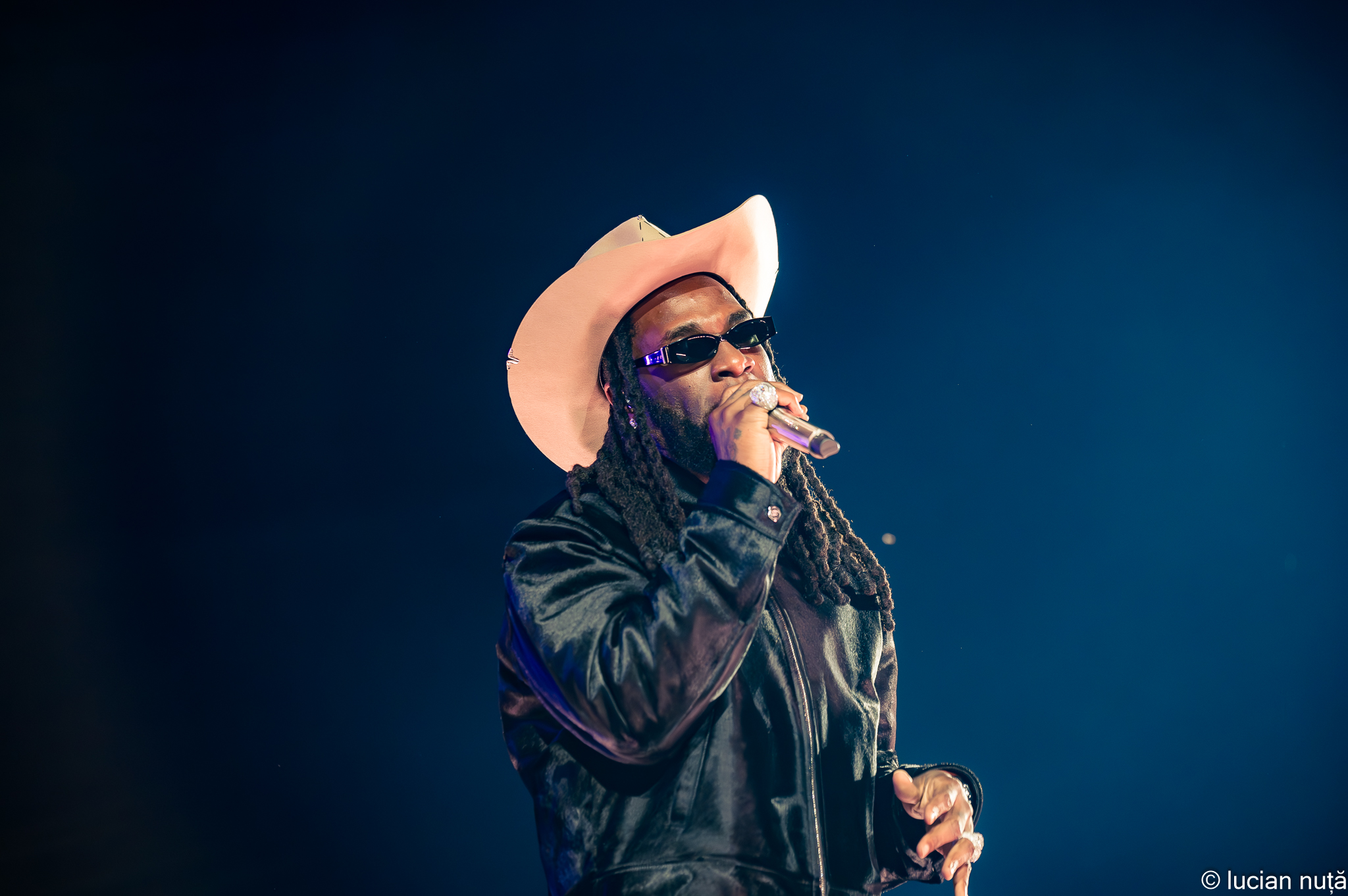 Burna Boy performing in a pale cowboy hat and sunglasses under blue stage light