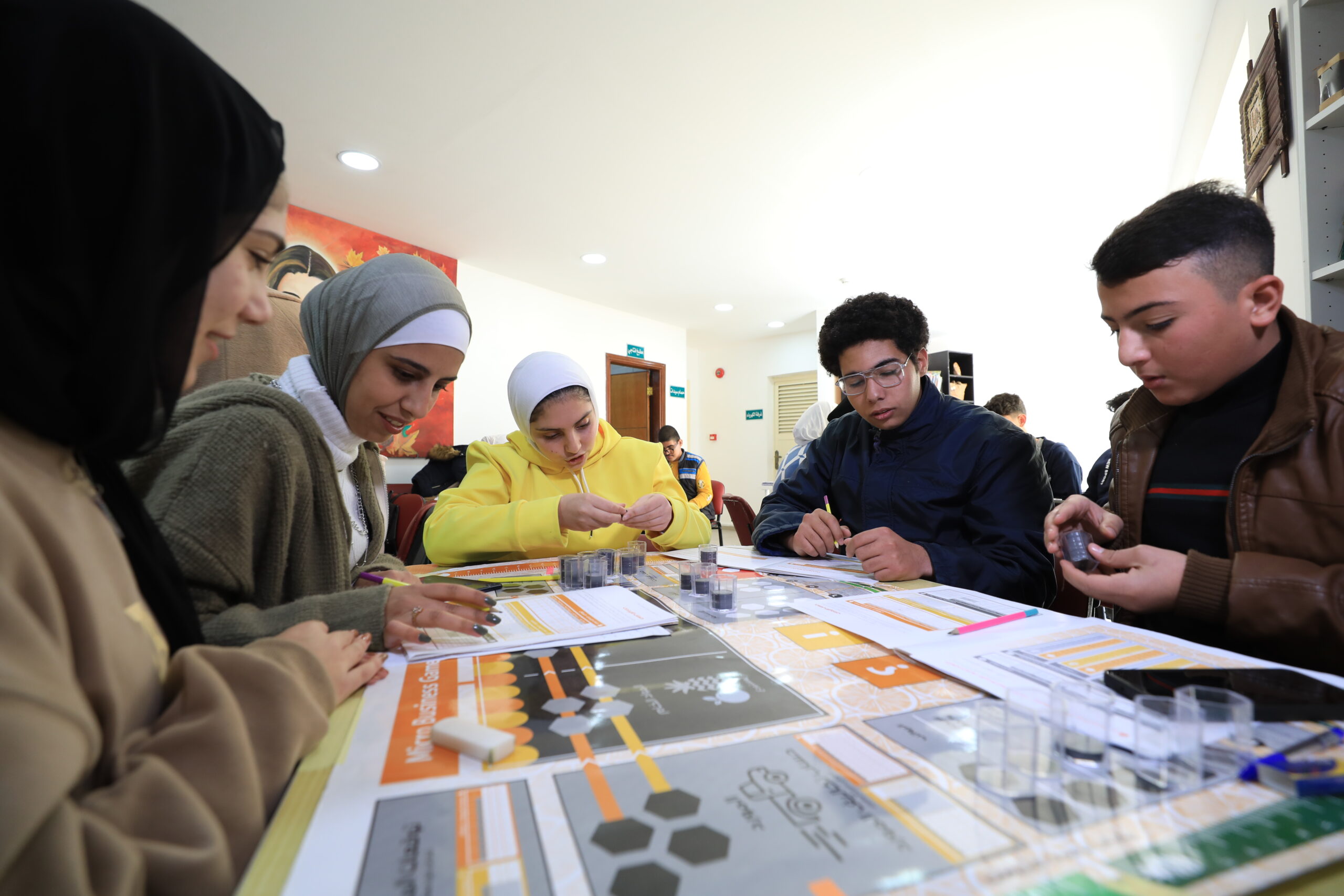 Young participants study worksheets and game pieces around a table during a BDC Micro Business Game session.