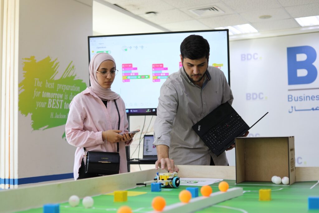 Two participants at a BDC Tariqi event demonstrate a small wheeled robot beside a laptop.