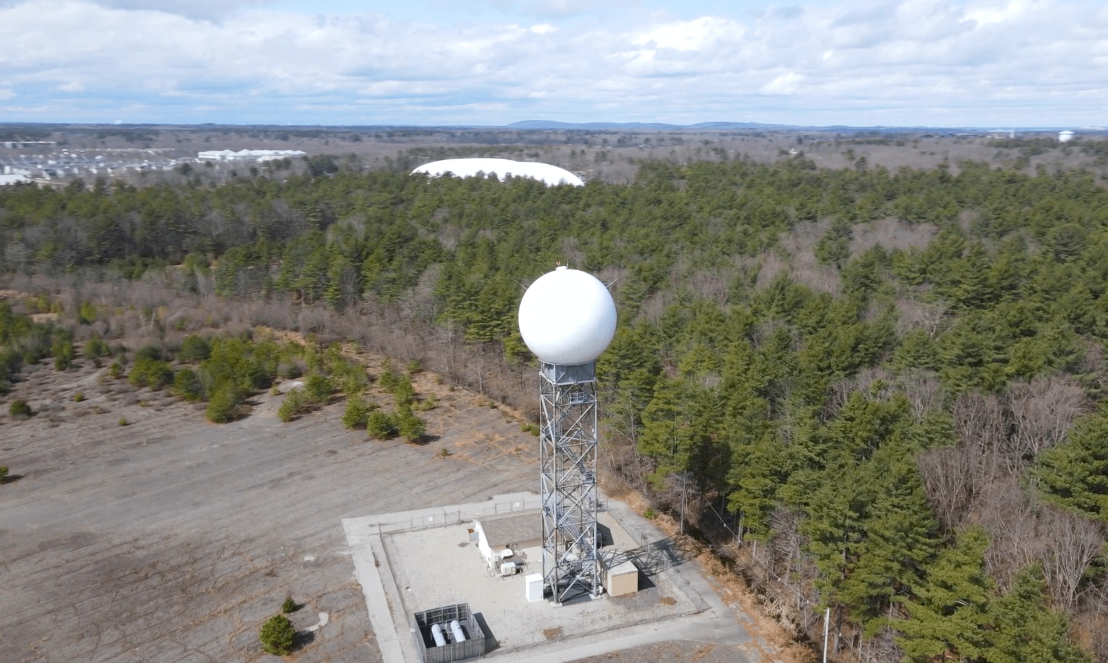 Radar installation with a white radome dome on a tower above a forest, an aerostat visible on the horizon