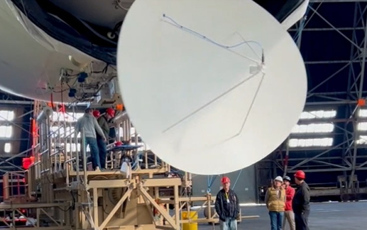 Workers in hard hats assembling a large white radar antenna dish inside a hangar