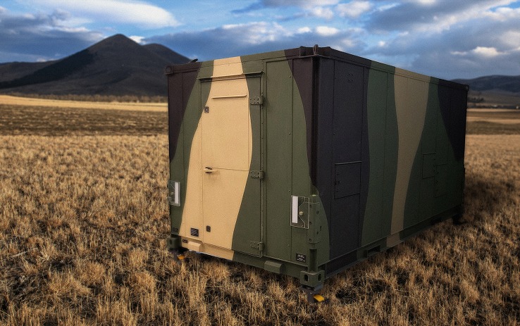 A camouflage-painted deployable equipment shelter sitting in an open field with hills behind it