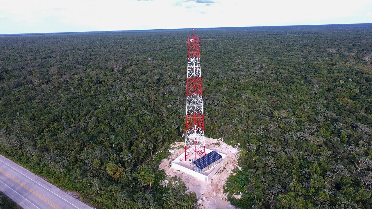 A red and white telecom tower beside solar panels, surrounded by forest