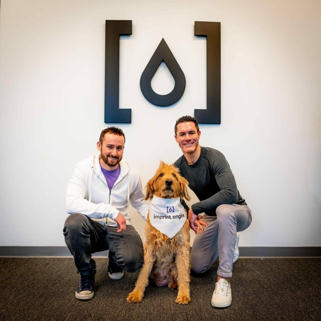 Caleb Gilbertson on the left and Travis Veit on the right, crouching beside a dog wearing an Imprint Engine bandana
