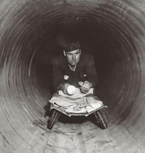 An archival Cal Water employee inside a large water main with a flashlight and plans