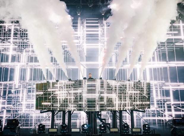 Calvin Harris performing behind a DJ console amid a geometric grid of white stage lights and smoke