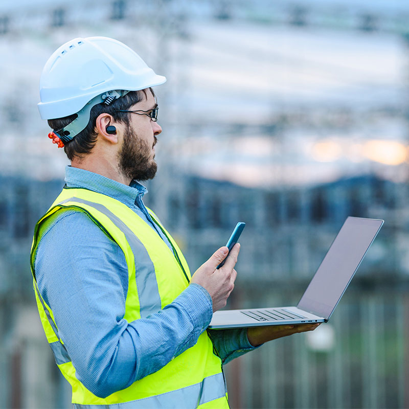 A worker wearing a hard hat and safety vest uses a laptop at an industrial site