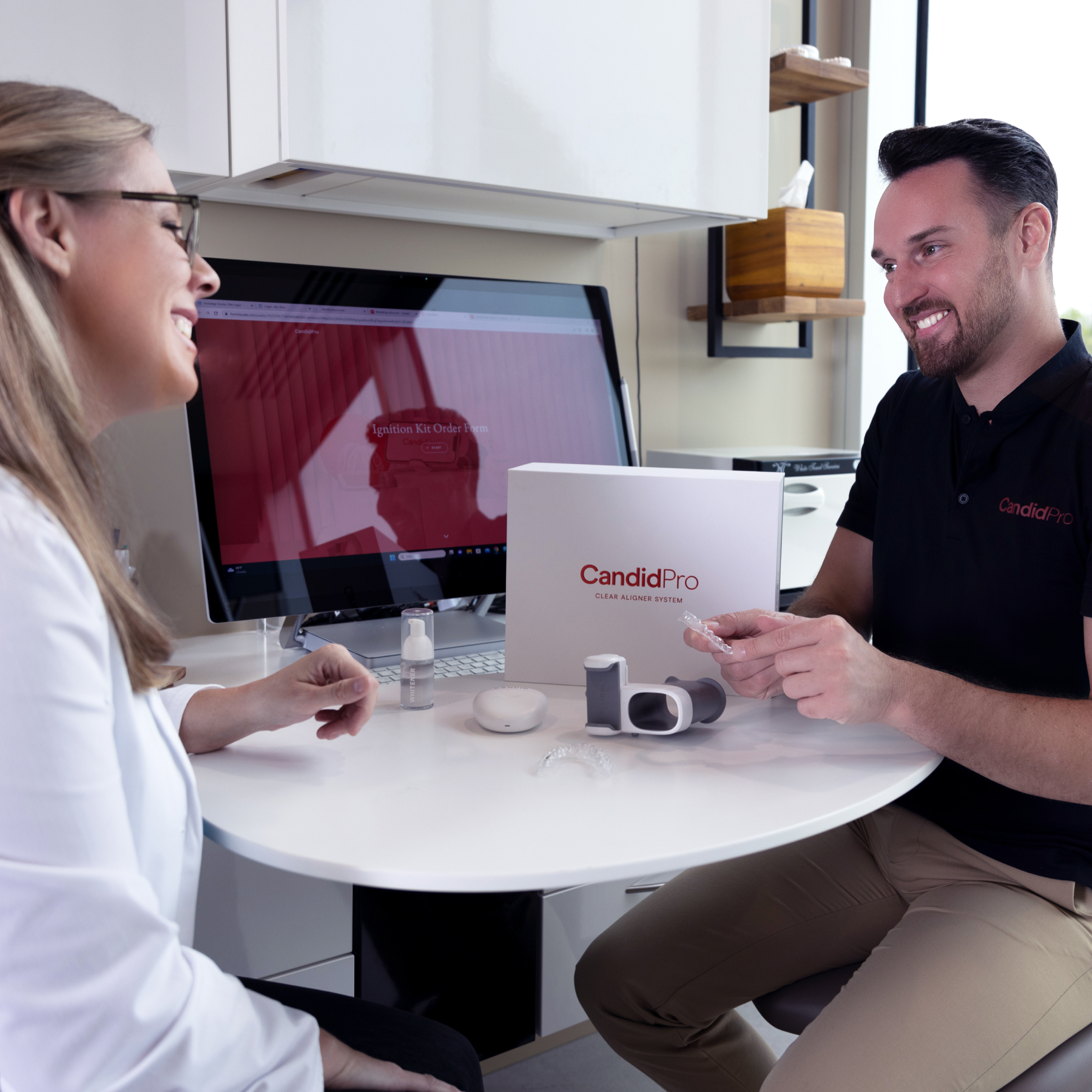 A dental professional and patient discuss CandidPro at a desk with aligner and monitoring equipment