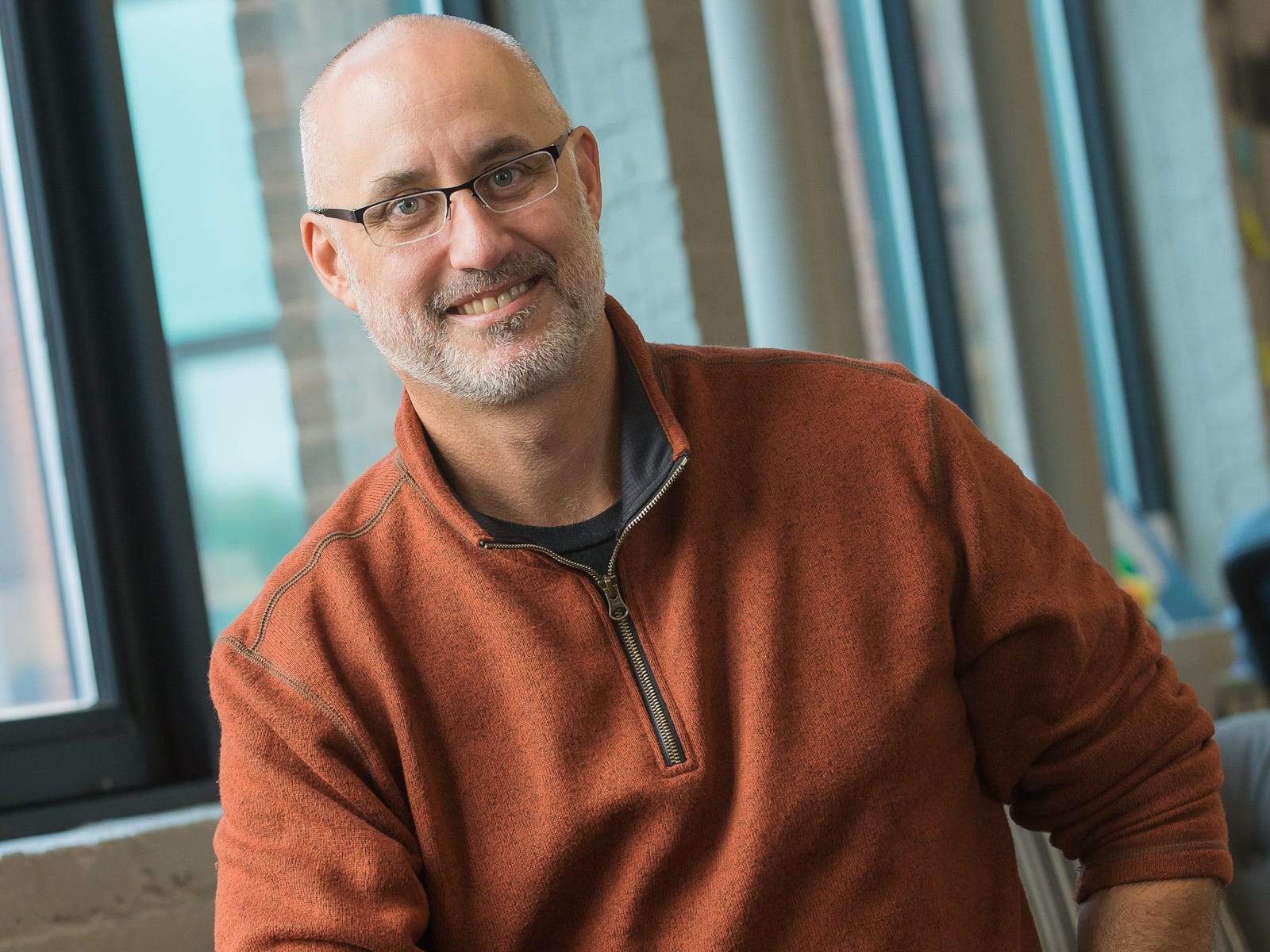 Carlos Cashman seated by office windows, wearing an orange quarter-zip sweater