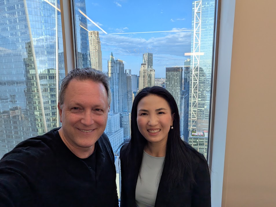 Ted Merz and Carmen Li smiling at Li’s New York office, with city buildings beyond the window