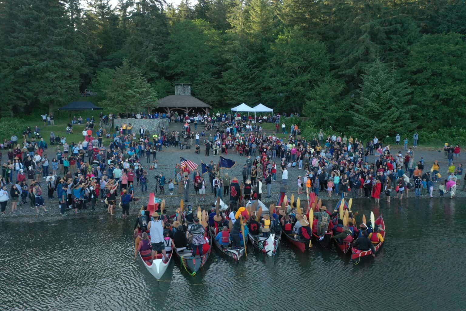 Colorful canoes approach a large community gathering on an Alaska shoreline, photographed from above