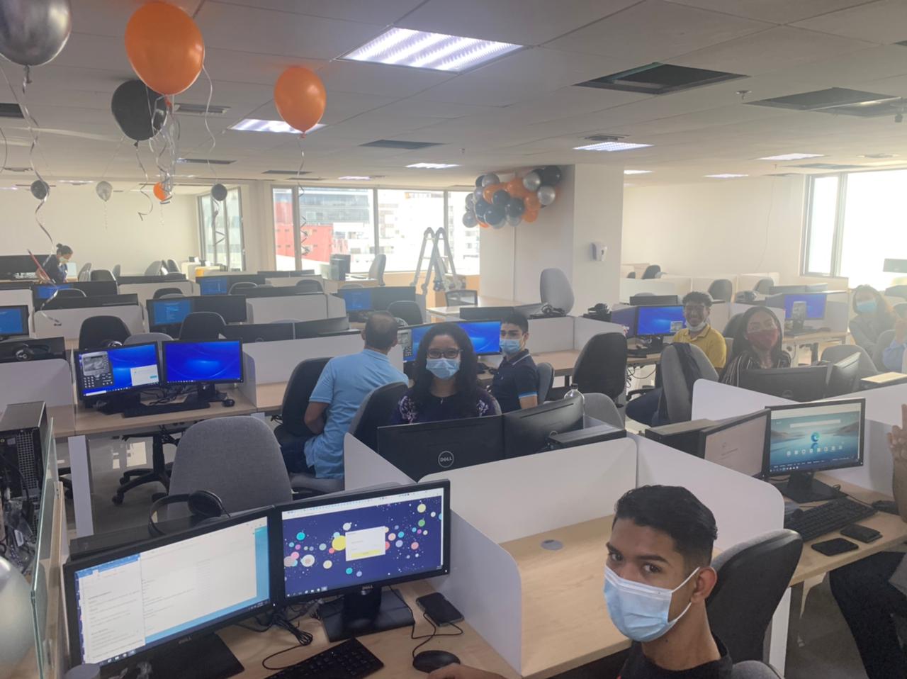 Employees at dual-monitor workstations beneath black and orange balloons in Center Source’s Barranquilla office