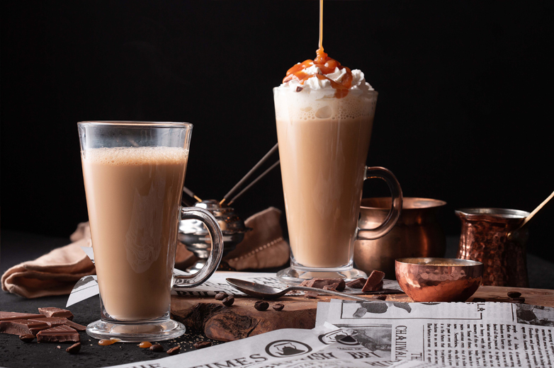 Two creamy Chaiiwala drinks served against a dark background with copper pots and spices