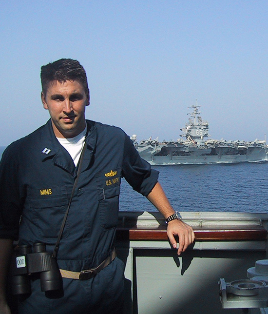 Chance Mims in Navy uniform aboard a ship, with an aircraft carrier behind him