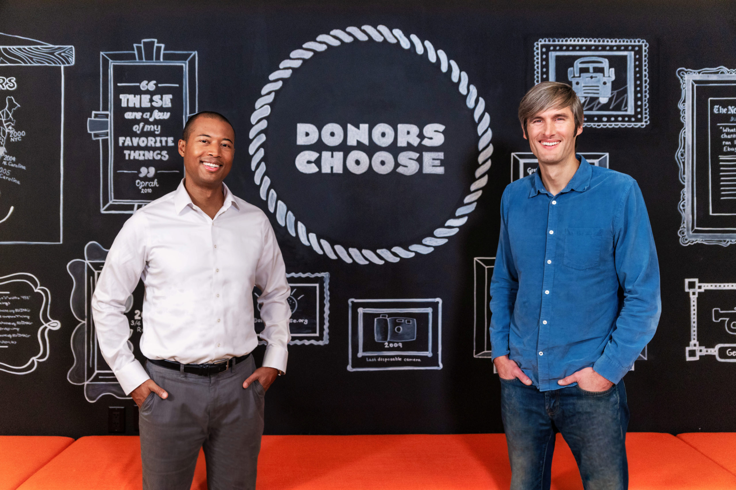 Alix Guerrier, left, and Charles Best, right, standing at DonorsChoose in front of a chalkboard wall.