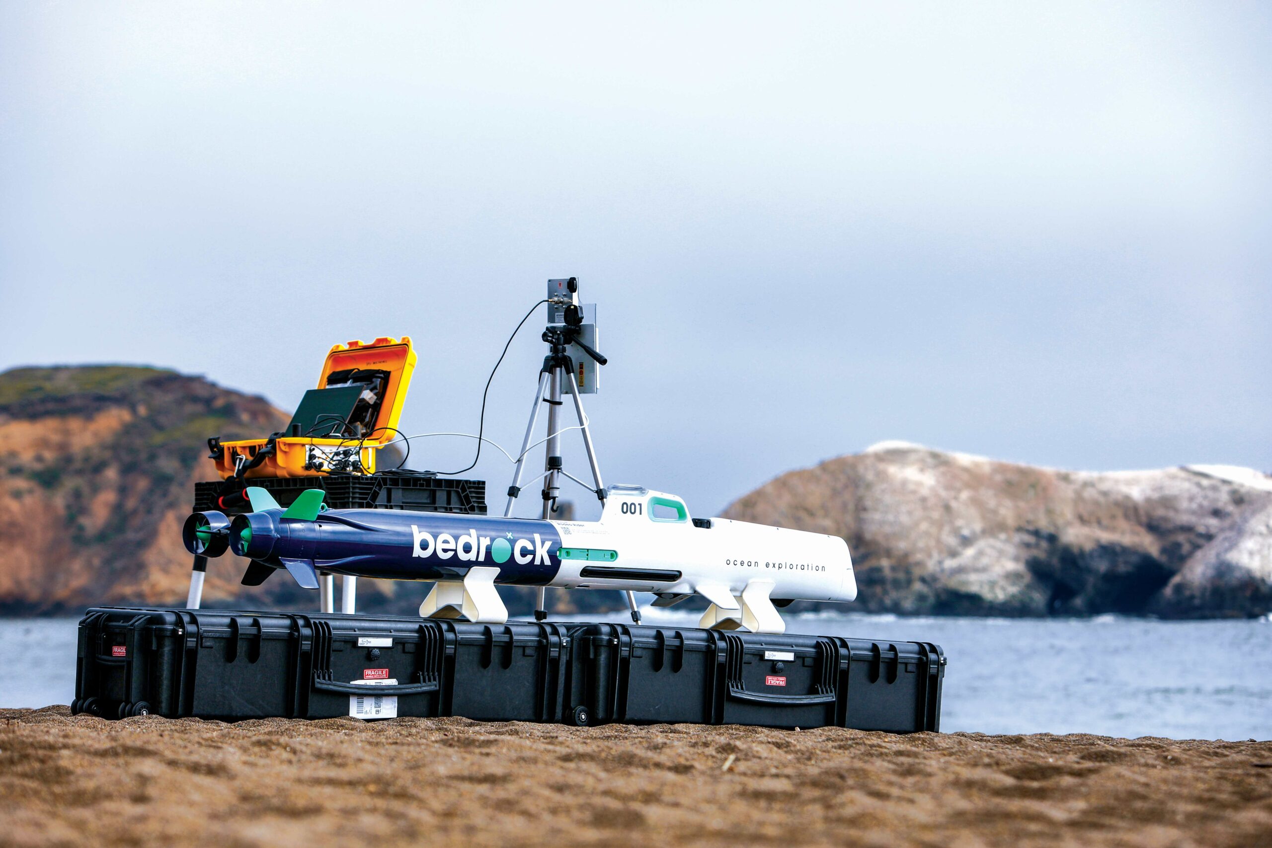 A white and navy Bedrock autonomous underwater vehicle resting on black transit cases beside the ocean