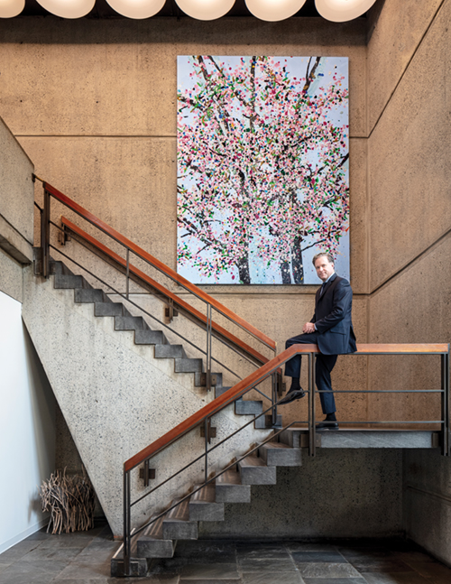 Charles Stewart sits on the staircase inside the Breuer building, beneath a colorful painting and circular ceiling lights.
