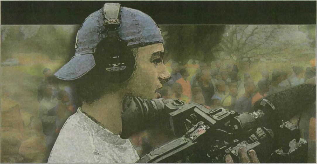 A young Charlie Ebersol wearing headphones and a backward cap while operating a video camera in South Africa