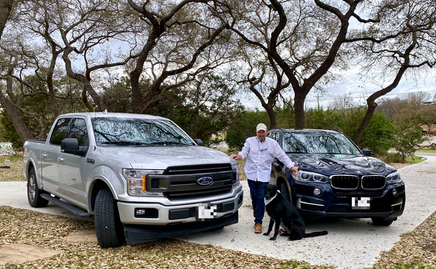 Chris Brown standing between a Ford pickup and a BMW SUV, with a black dog beside him