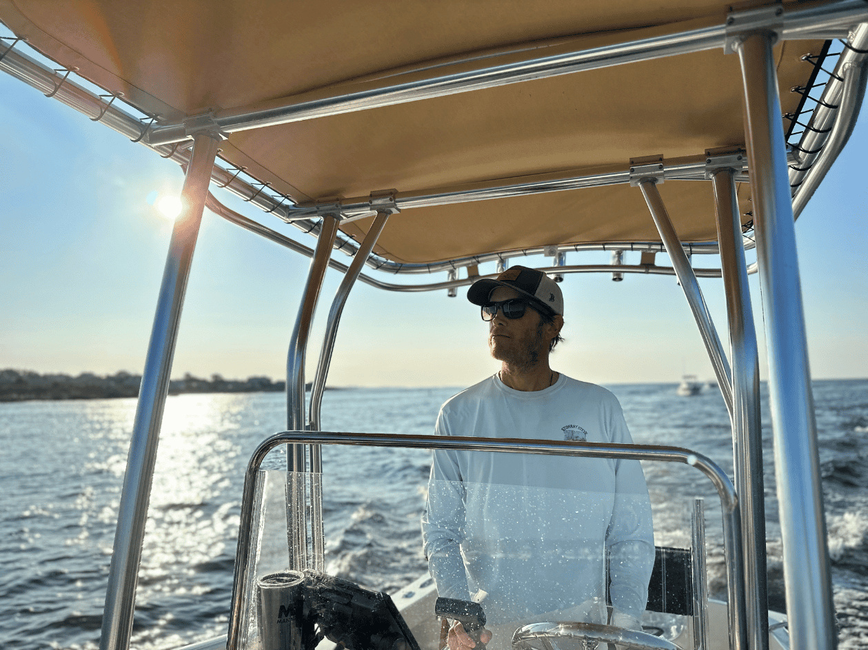 Chris Gosline wearing a cap and sunglasses at the helm of a boat, with sunlit water behind him