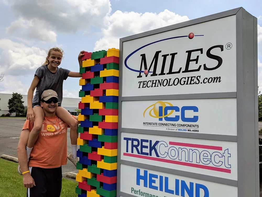 Chris Miles carrying a child on his shoulders beside the Miles Technologies sign and a column of colorful oversized blocks