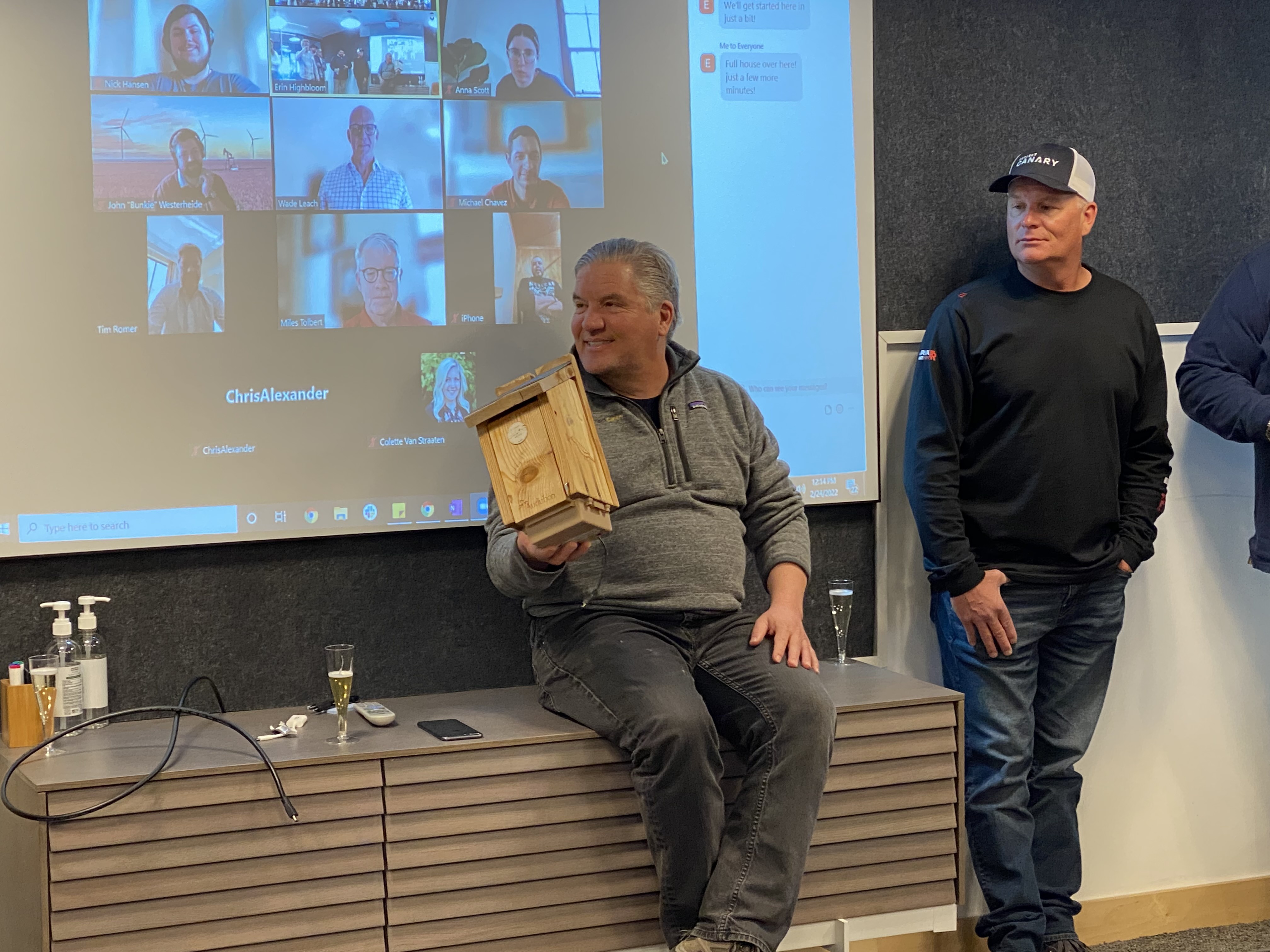 Chris Romer holds a wooden monitoring device at a Project Canary team meeting, with colleagues on a video call behind him.