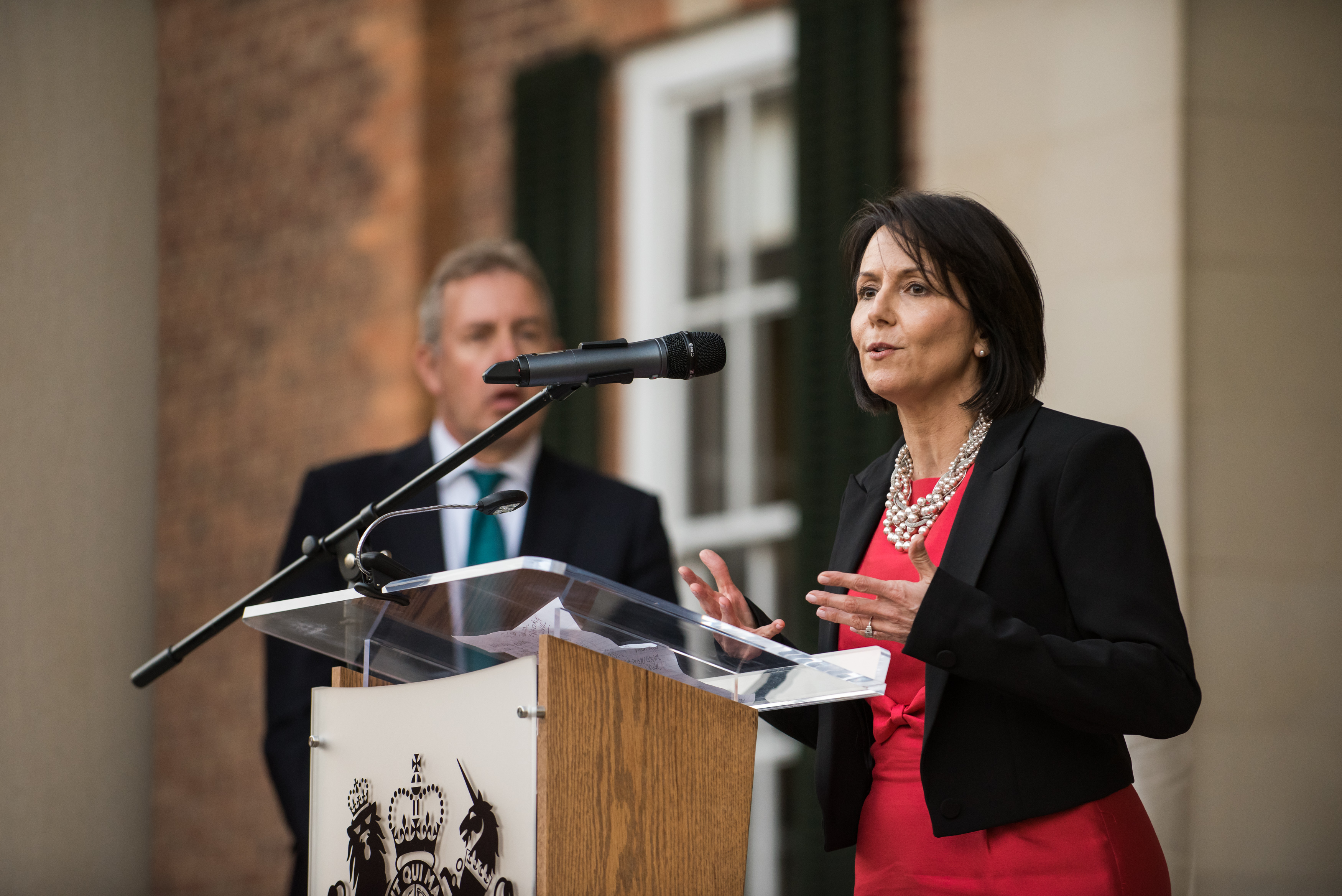 Christa Gyori speaking into a microphone at a lectern, wearing a red dress and black jacket.